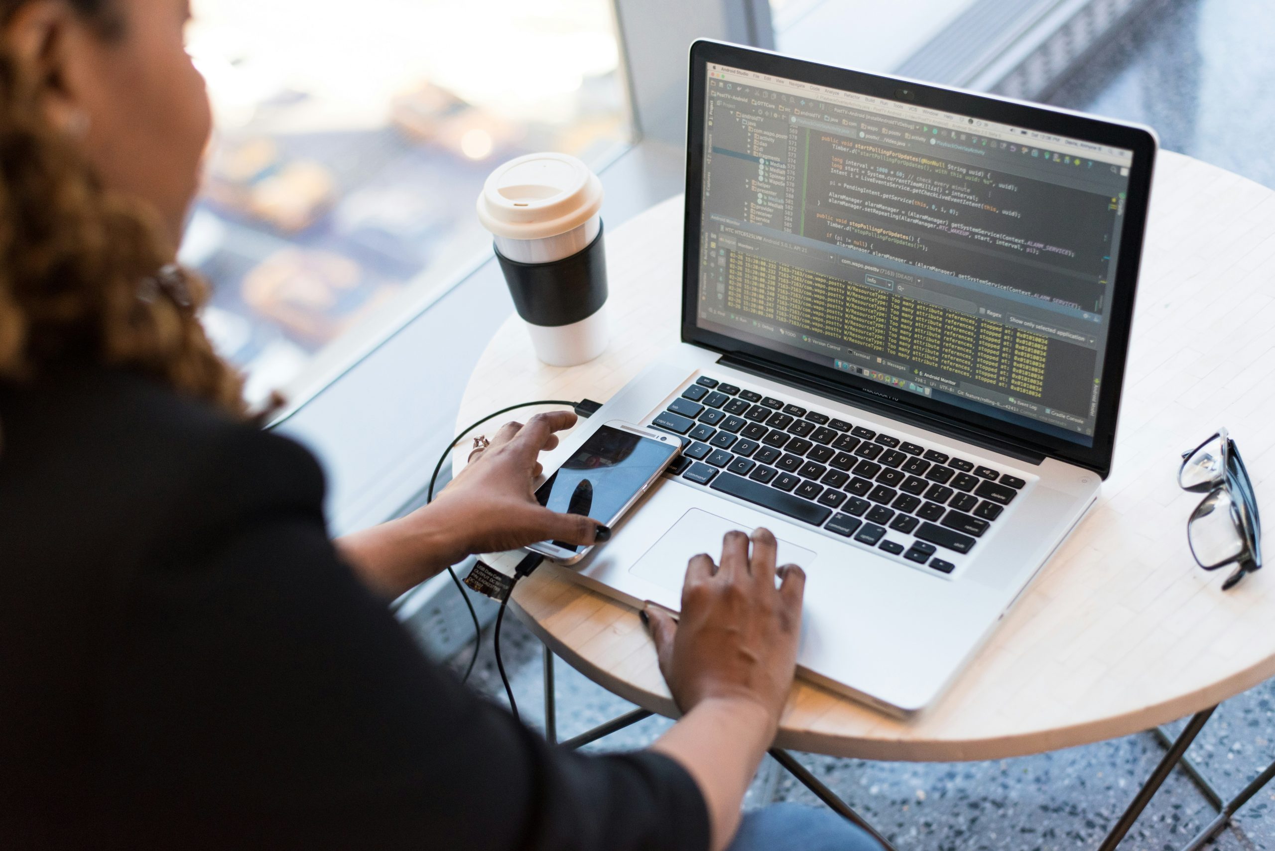 Black woman entrepreneur coding on a laptop with her right hand, a coffee on her left and a cell phone in her left hand.
